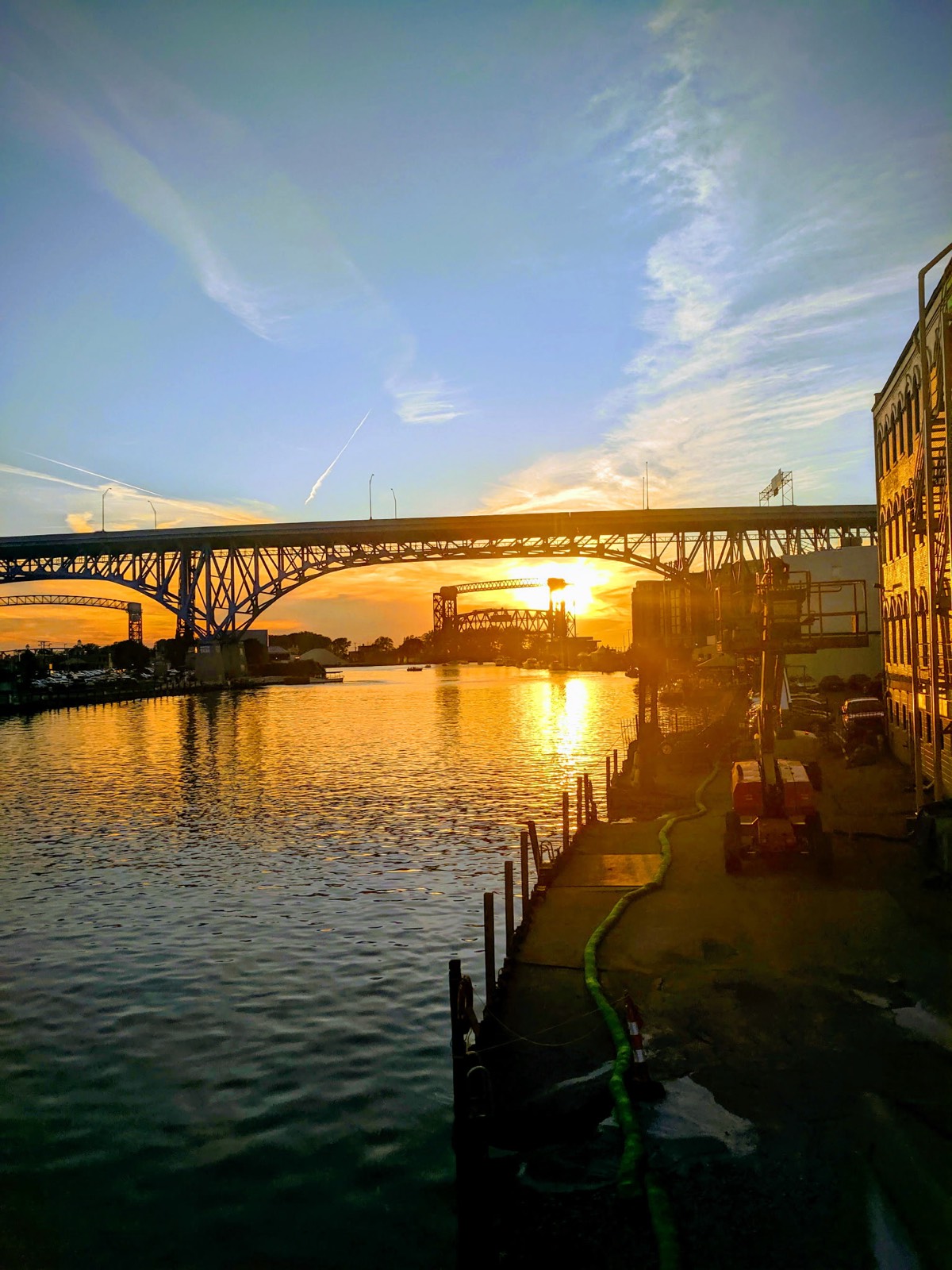 The sun setting down the Cuyahoga River, seen from the Flats, with a high steel arch bridge crossing the frame and a lift bridge silhouetted behind it.