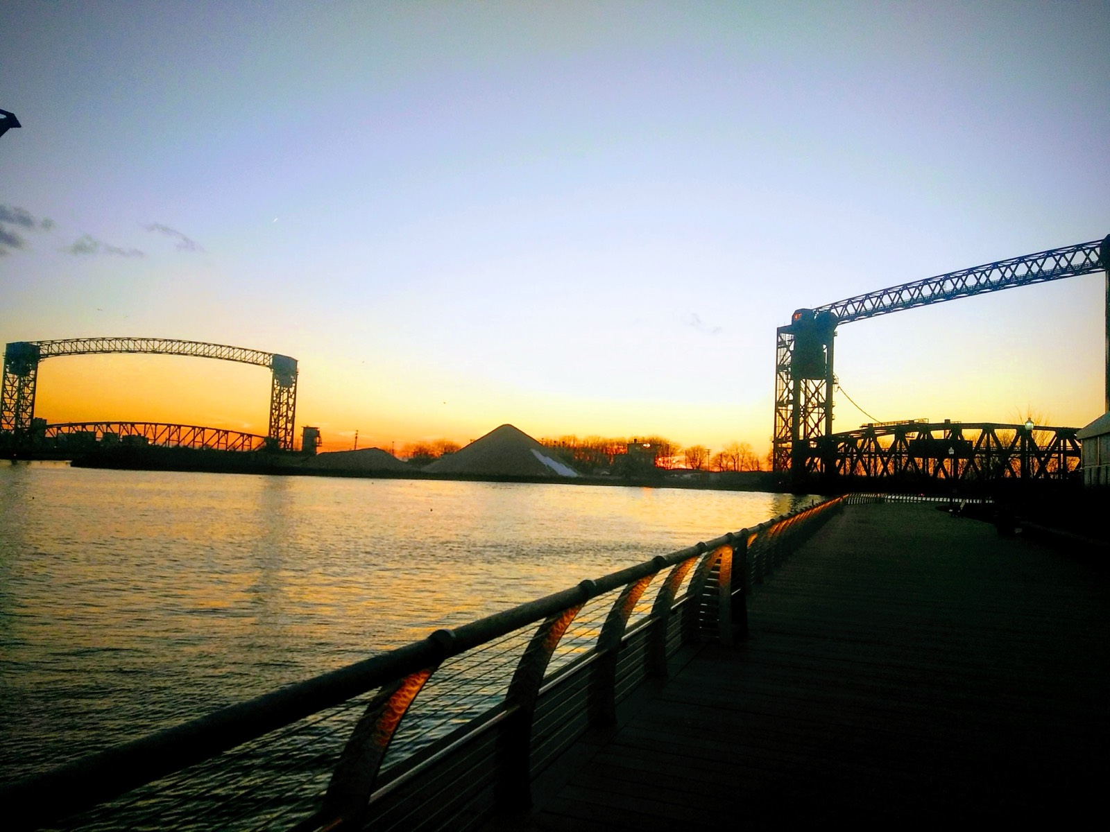 Two vertical-lift railroad bridges standing open on either side of the Cuyahoga at dusk, seen from a riverfront boardwalk.