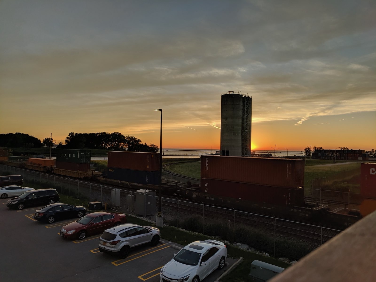 Sun dropping into Lake Erie behind a concrete silo, with a line of stacked shipping containers on a freight train in the foreground.